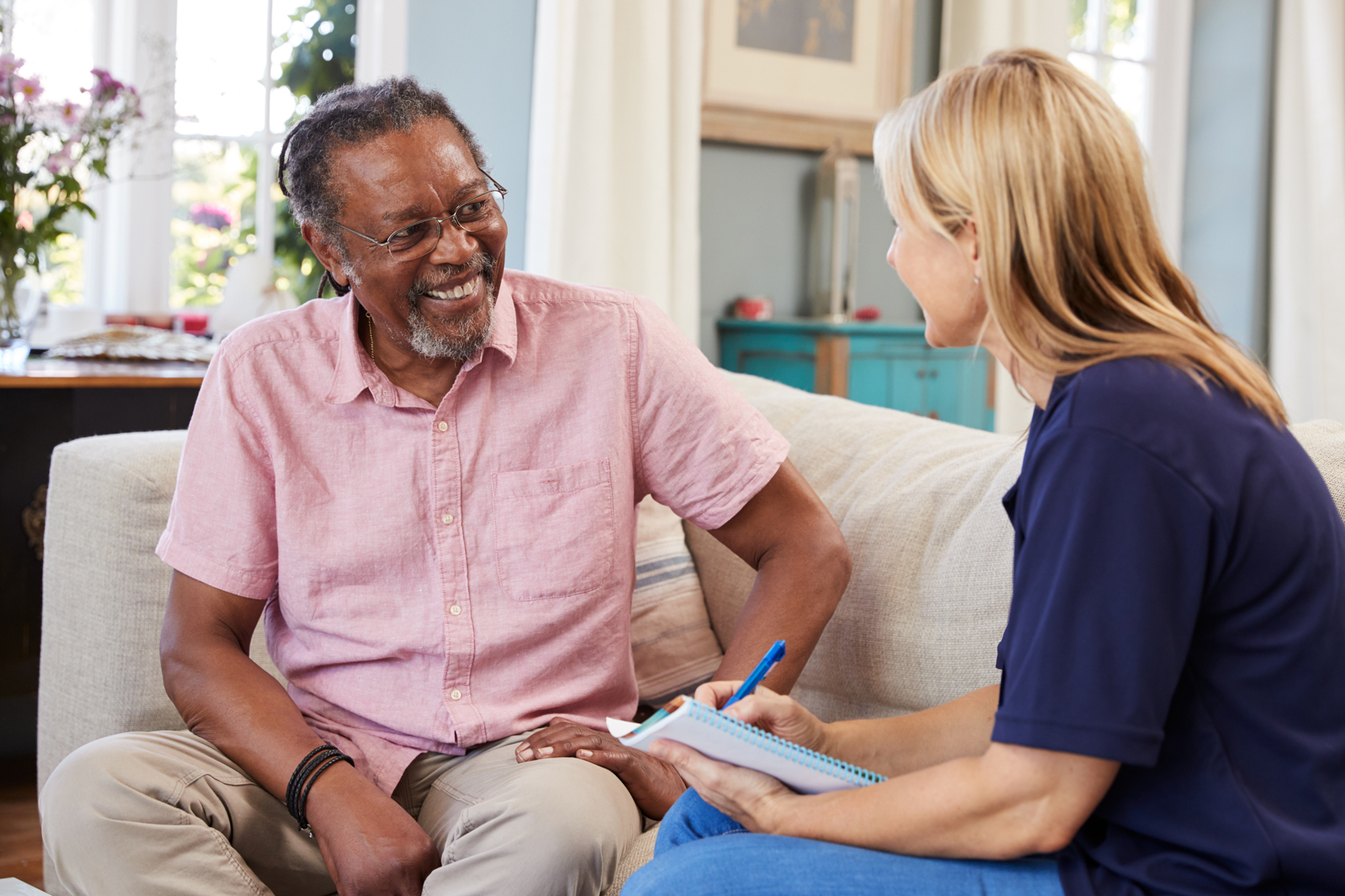 An older adult Black man sits on a sofa with an aide who is holding a clipboard with forms.