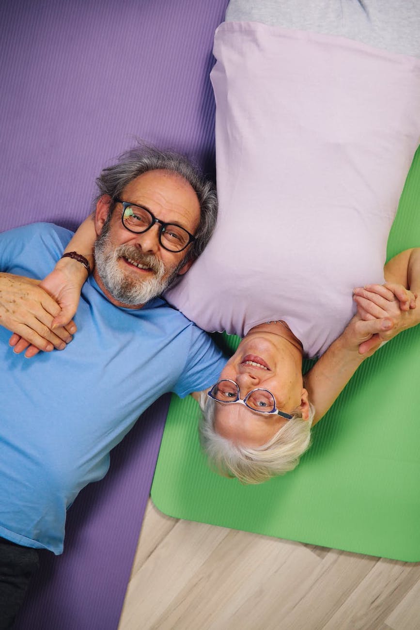 An older adult man and older adult woman lay on yoga mats and embraces.