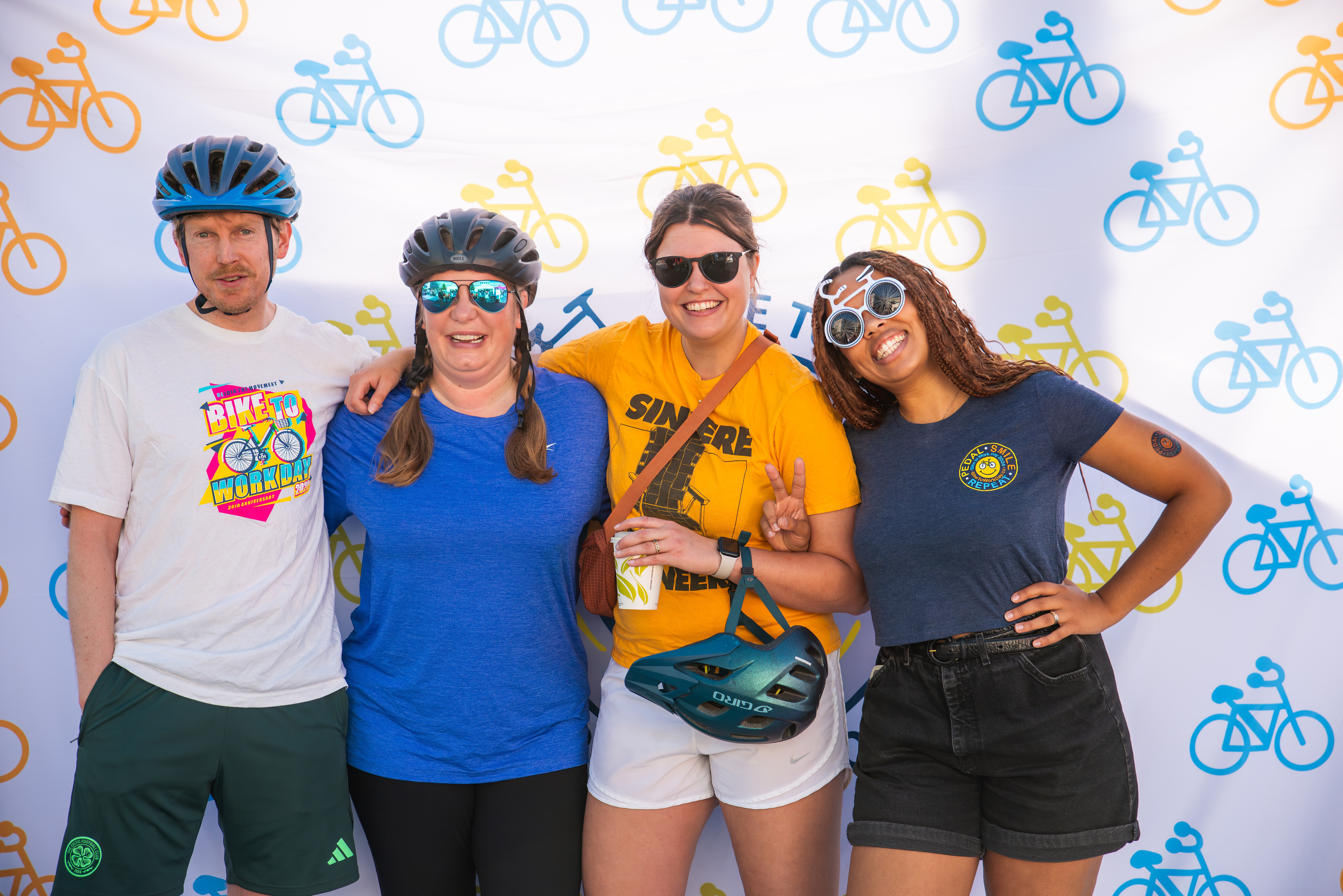Four people smile in front of a Bike to Work Day backdrop in t-shirts, shorts, sunglasses and bicycle helmets.