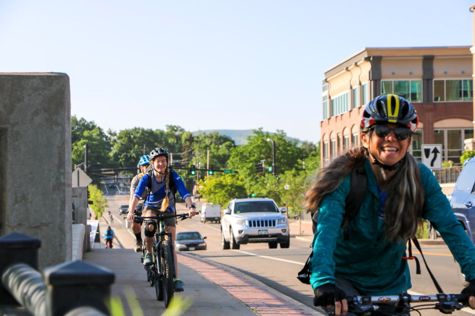A commuter riding a bicycle at the forefront of the image with two additional commuters riding bicycles in the background. Vibrant, greens foliage, commercial buildings and cars on the road are in the background of the image.
