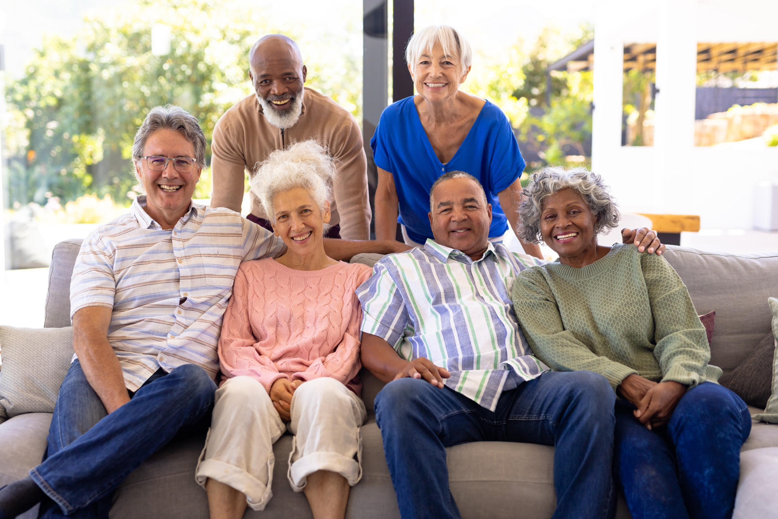 A racially diverse group of four older adults sit on a couch from left to right of man, woman, man woman. Two older adults stand behind the couch in the order of man and woman.