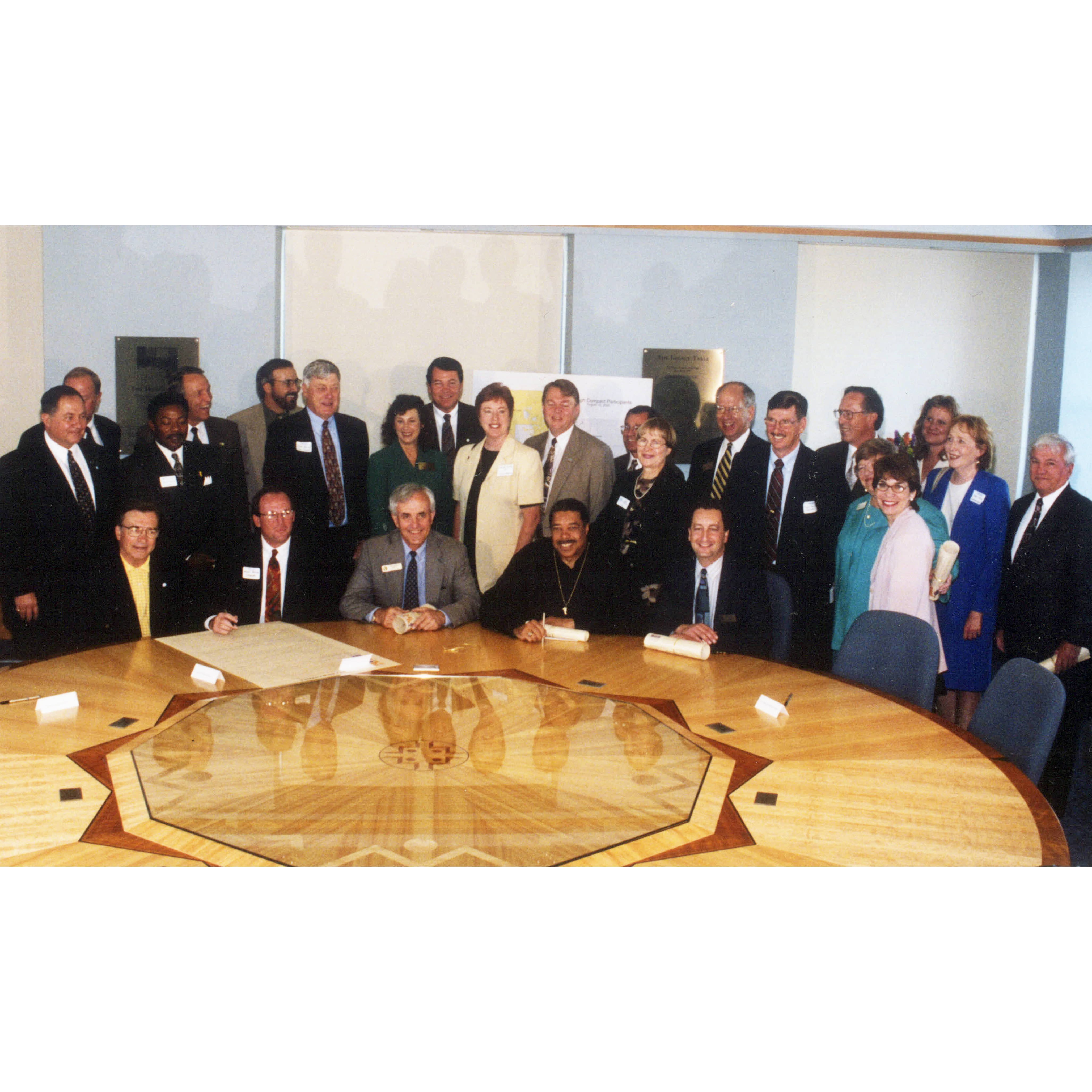 Representatives from five communities and 25 municipalities stand and sit before a large table during the Mile High Compact signing ceremony on August 10, 2000.