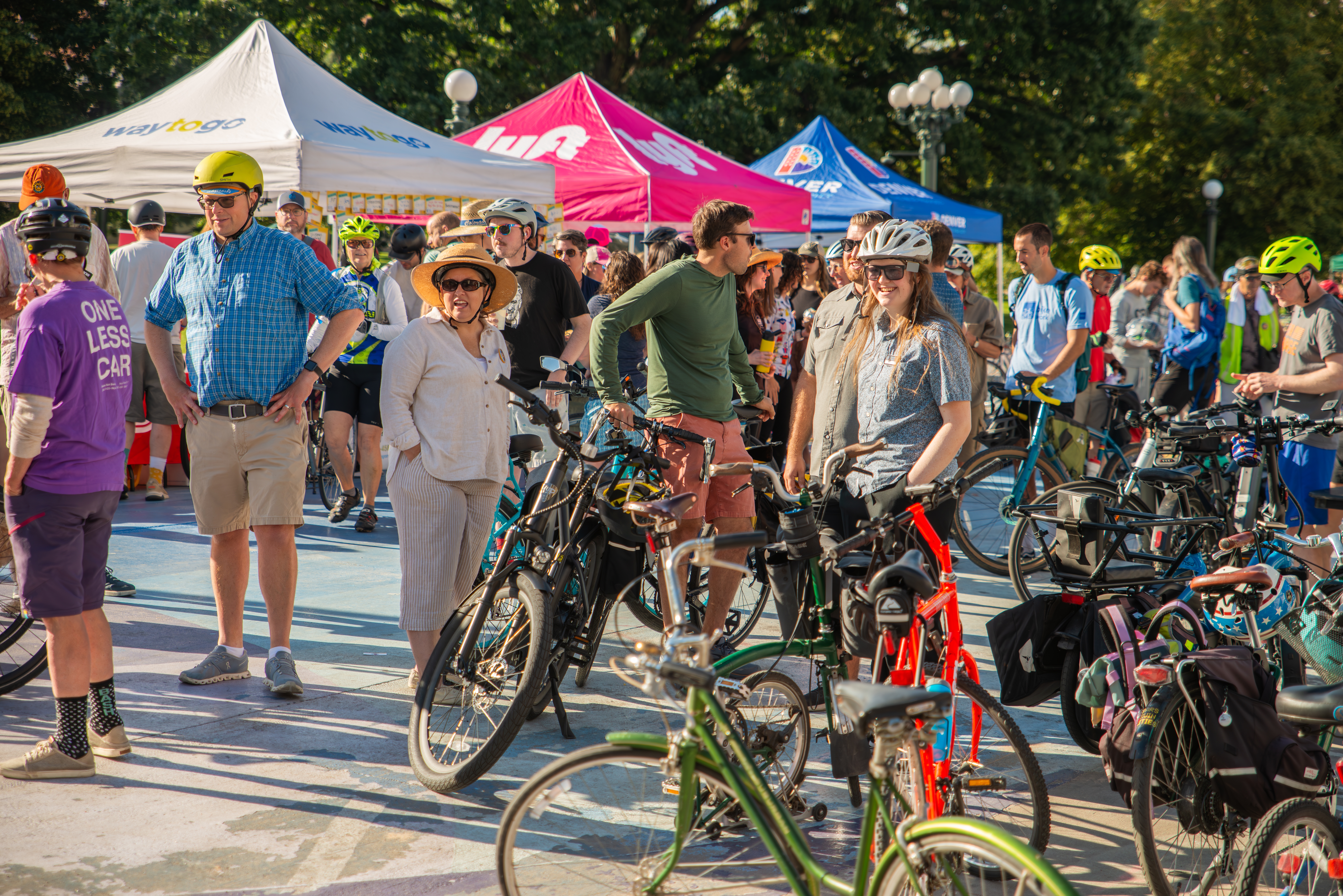 A group of people standing in together with their bicycle helmets and gear on. In the foreground of the image is a group of parked bicycles and in the background is booths set up for Bike to Work Day 2024.
