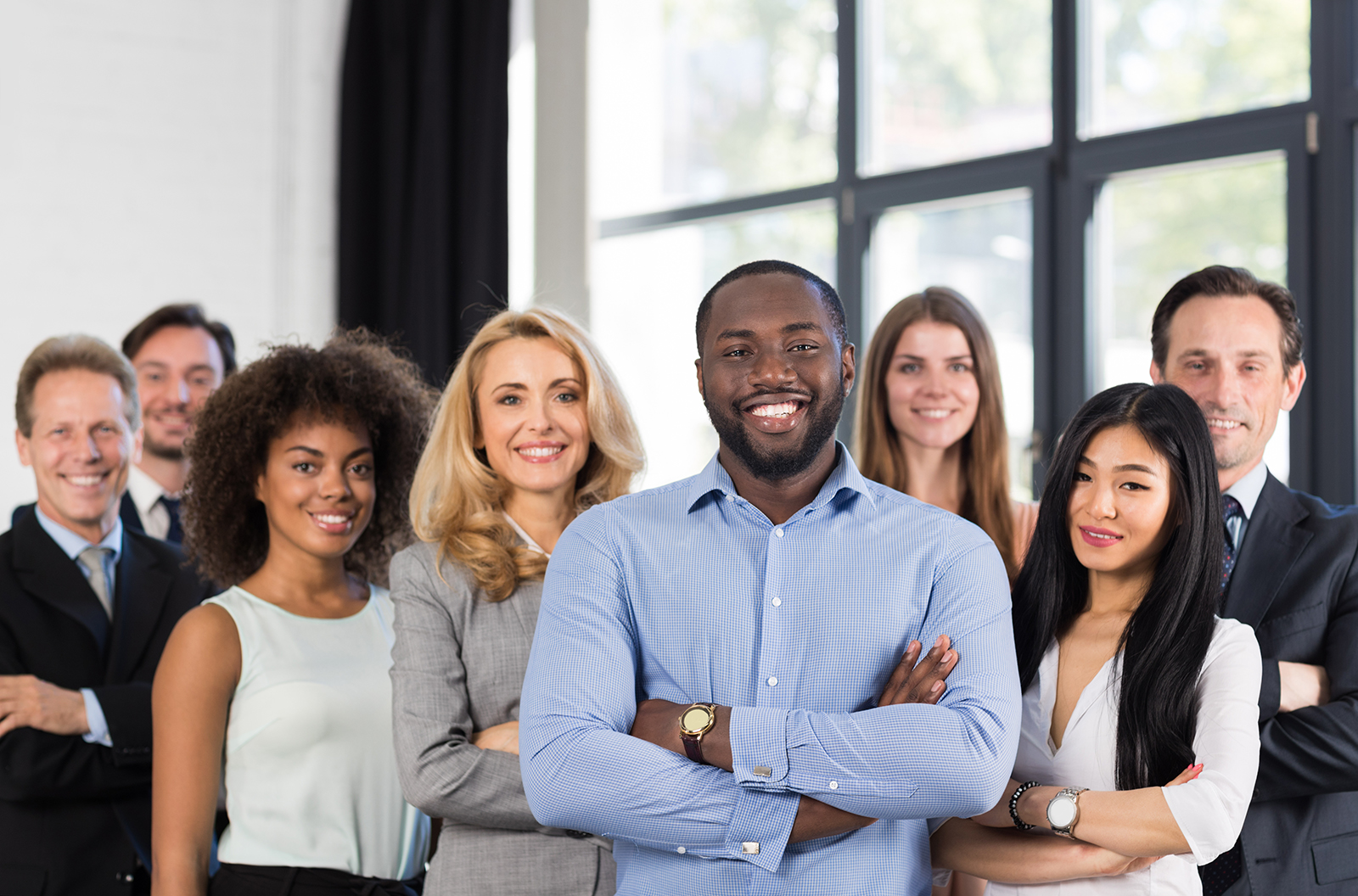 Several people of various races and ethnicities, all wearing business attire, stand smiling in front of a large office window.