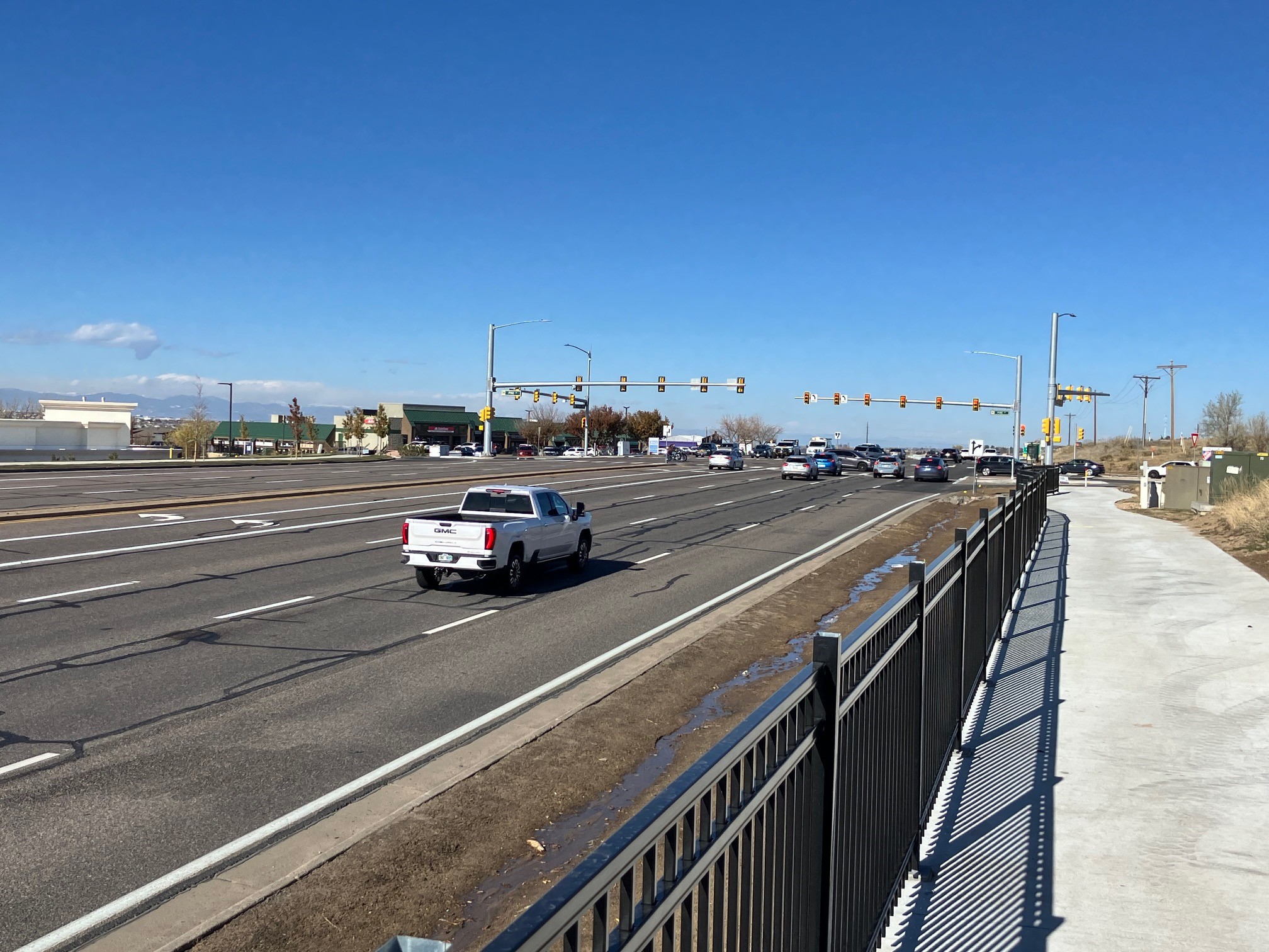 A concrete path with a black metal fence running alongside it sits next to a six-lane road on which several cars and trucks are traveling.