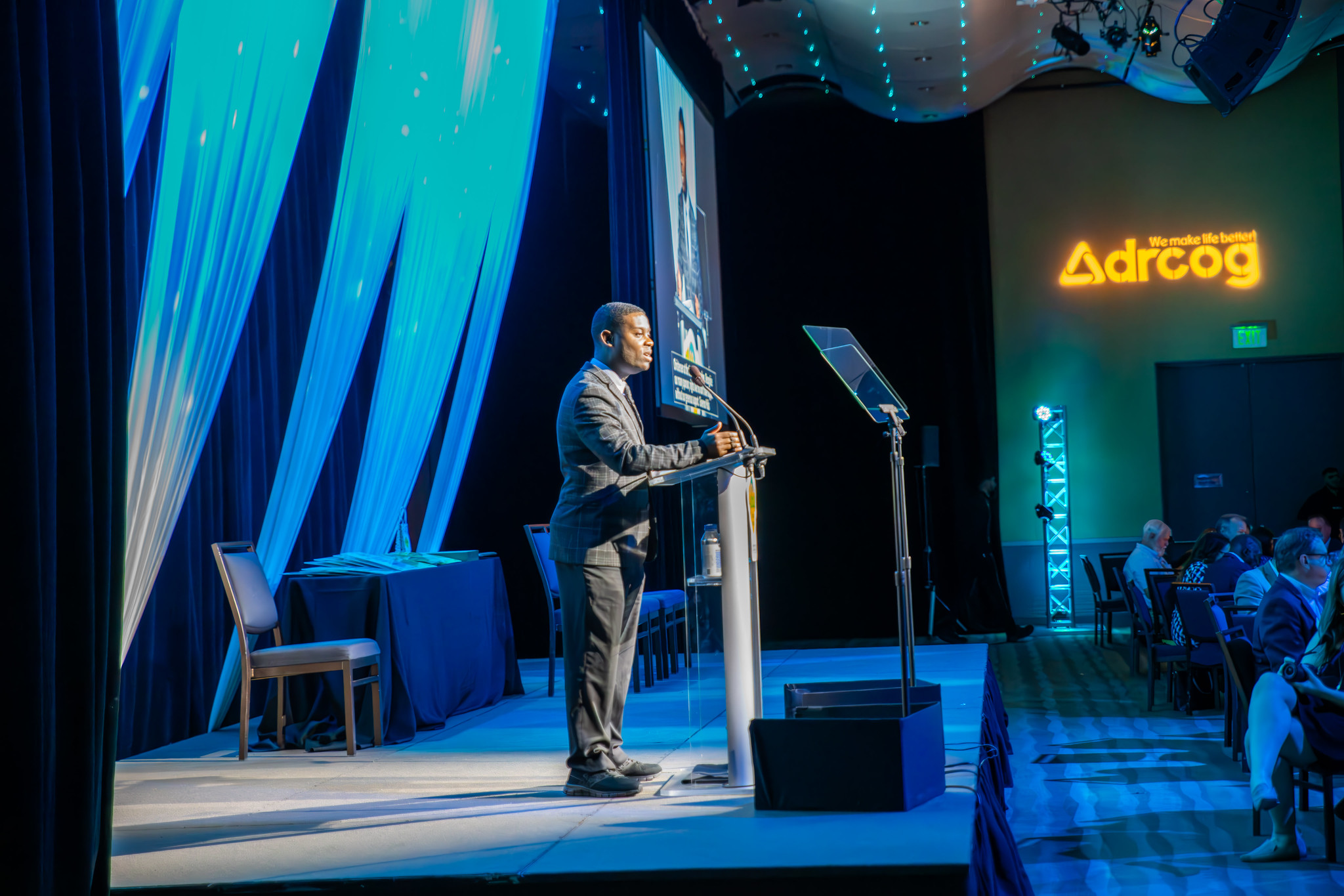 A Black man stands at lectern in a dimly lit ballroom. 