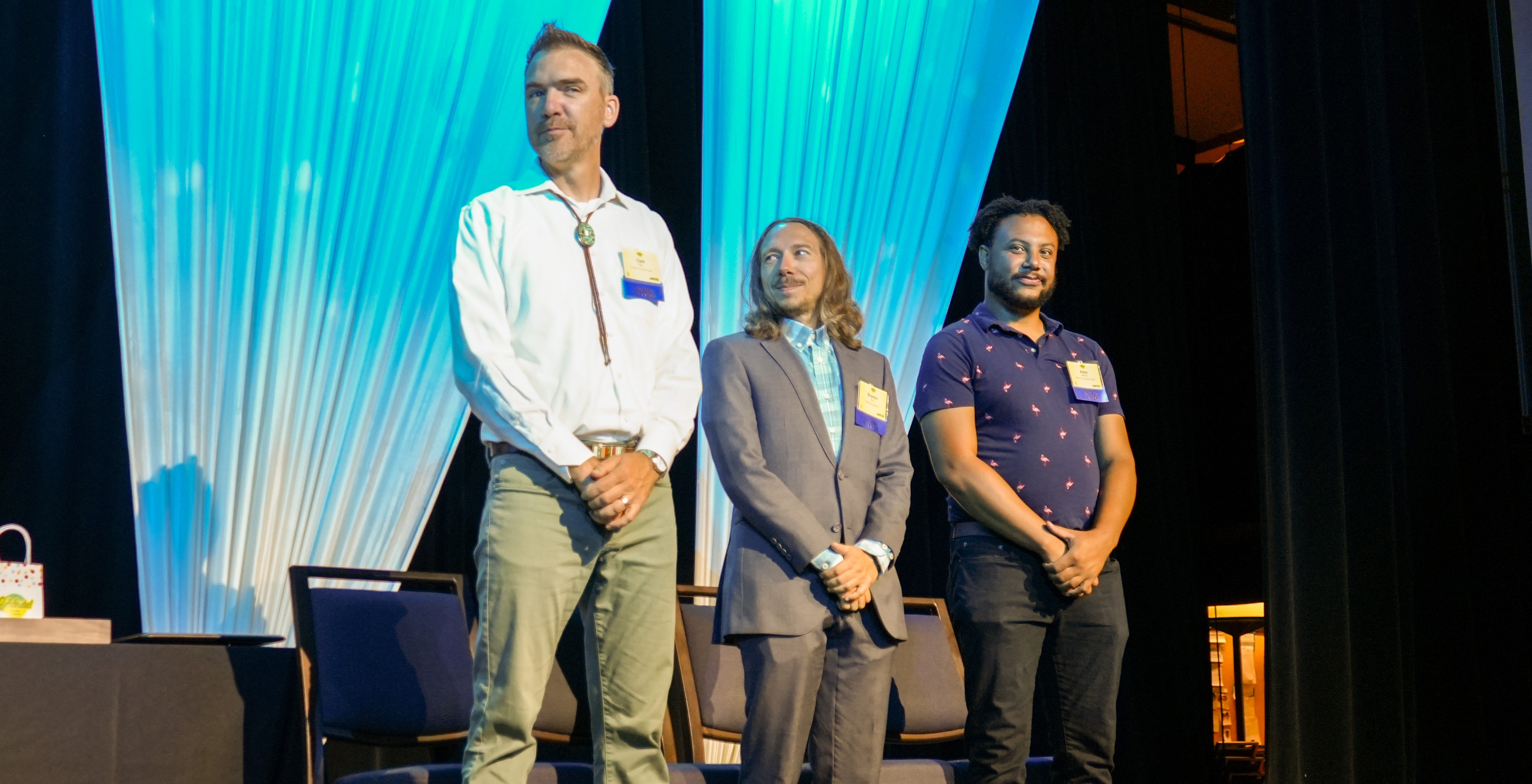 Three people stand on a stage with a blue curtain.