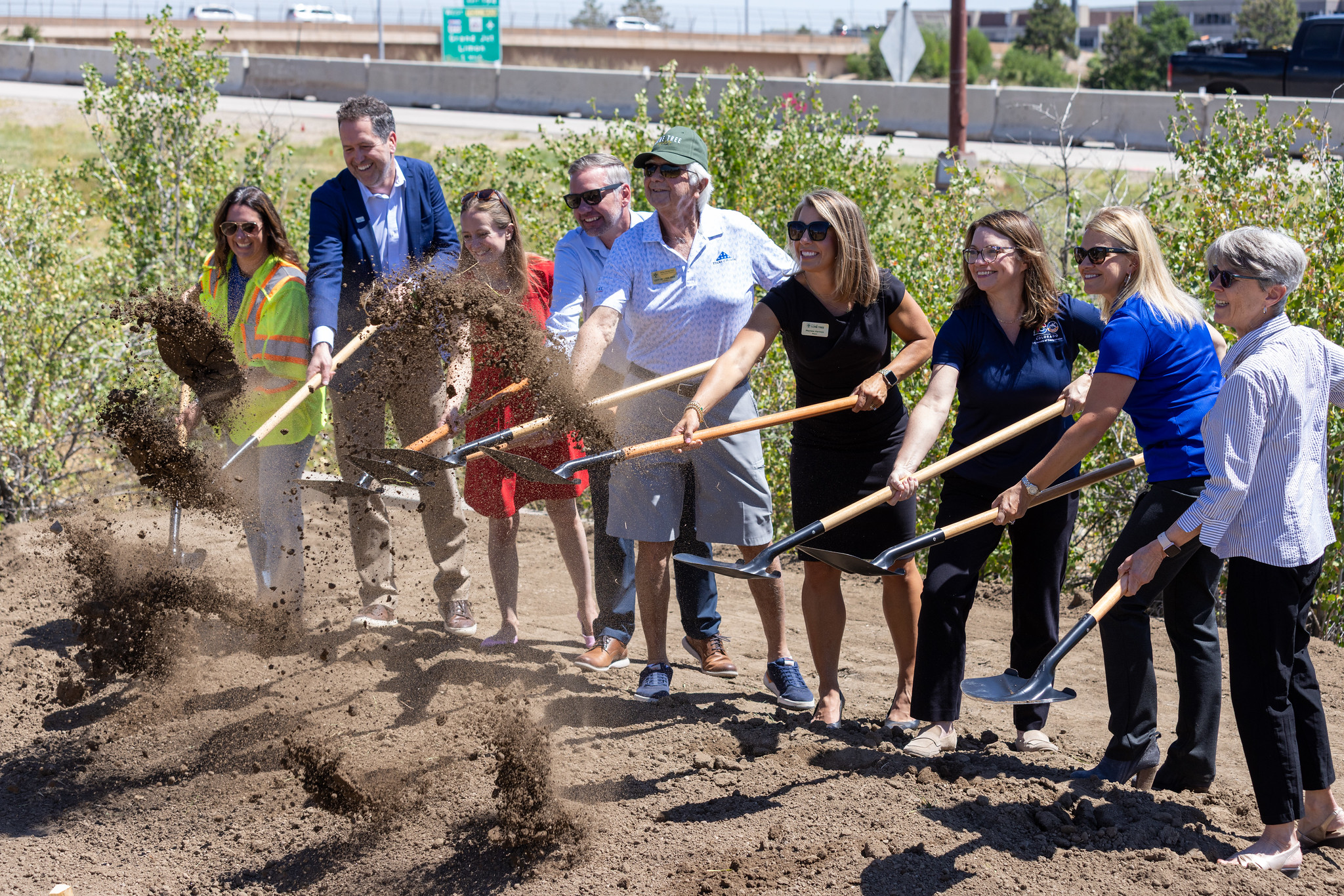 A group of nine people holding shovels stand side-by-side in a line and toss dirt in the air as part of a ground breaking.