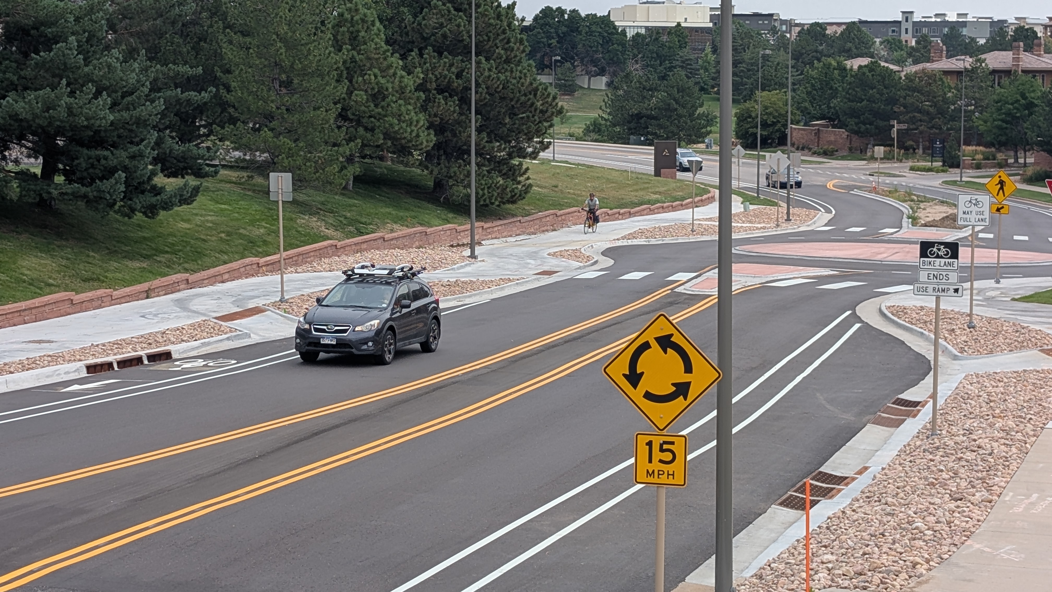 A black car drives down a mostly empty road with bike lanes and new sidewalks on each side. A small roundabout can be seen in the background.
