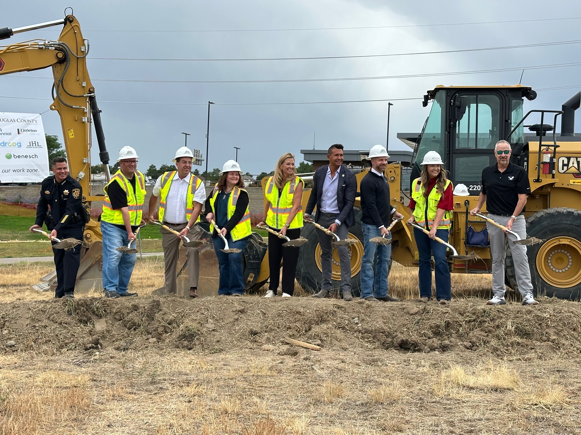 Several people, most of whom are wearing hard hats and safety vests, stand next to a backhoe, smiling and holding shovels full of dirt.