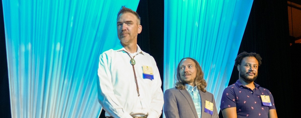 Three people stand on a stage with a blue curtain.