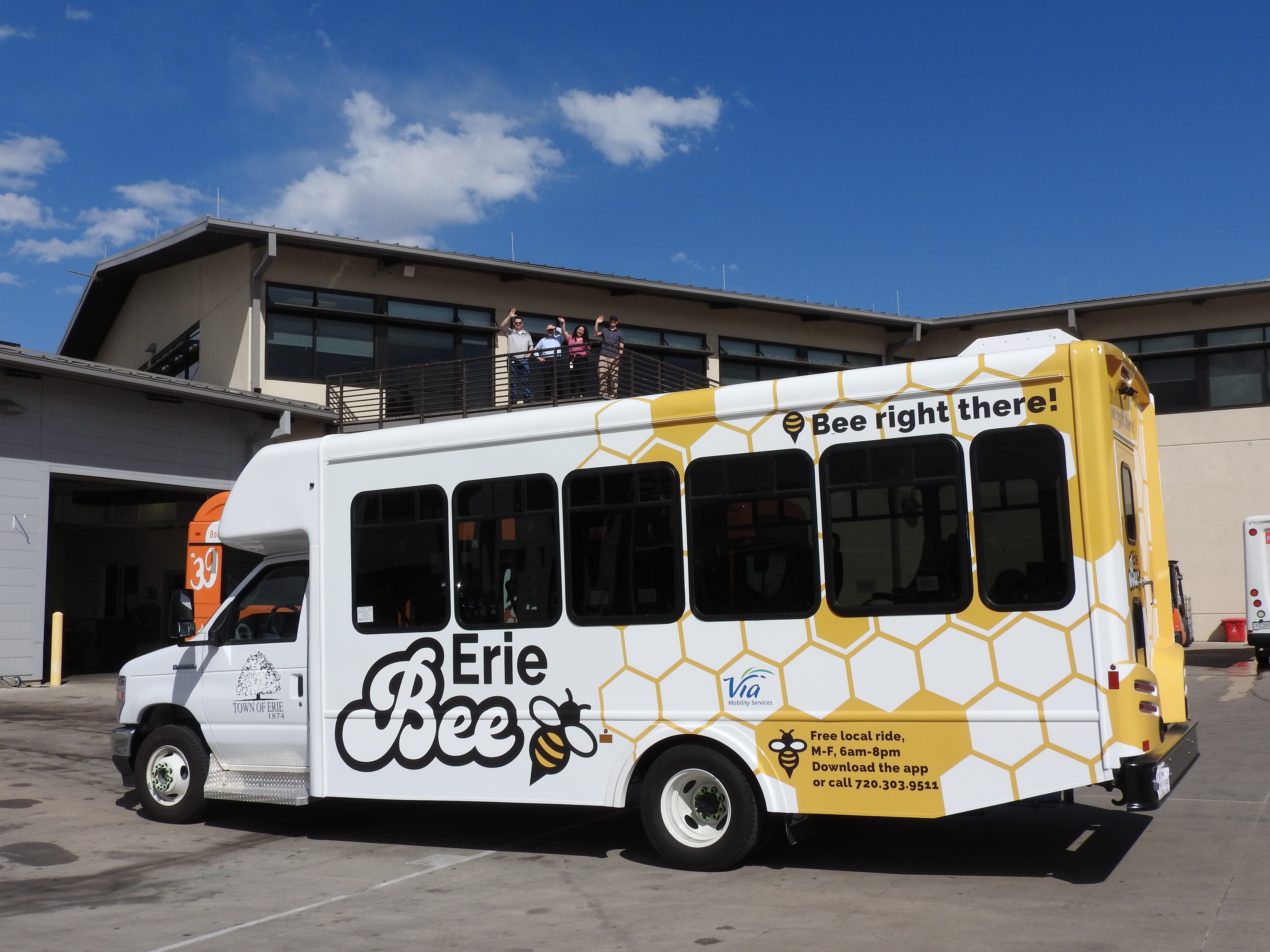 Erie Bee shuttle parked outside a building, with a yellow honeycomb design and “Bee right there!” slogan, while several people stand on a balcony above and wave.
