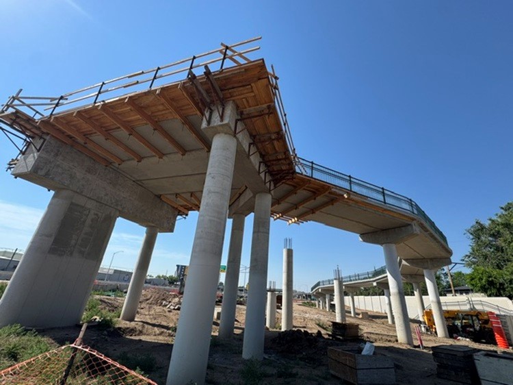 The underside of a tall bridge under construction. A finished ramp leads to a large platform with wooden supports still in place.