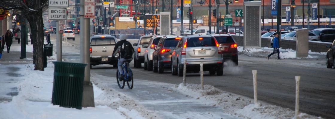 A line of cars waiting at a red light in Downtown Denver. A person riding a bike is approaching the red light. There is snow on the ground and commercial buildings in the background.