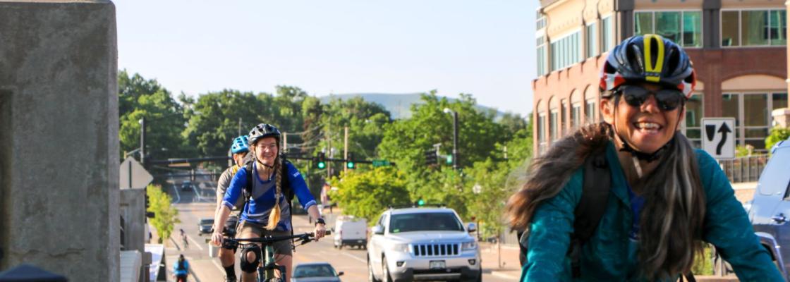 A commuter riding a bicycle at the forefront of the image with two additional commuters riding bicycles in the background. Vibrant, greens foliage, commercial buildings and cars on the road are in the background of the image.