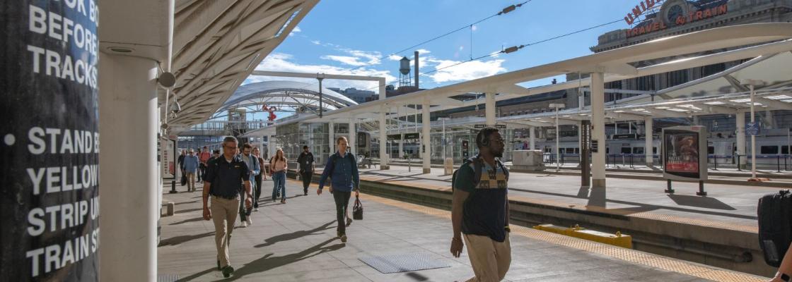 A group of commuters walking past the rail lines at Union Station in downtown Denver. Union Station's sign in in the background and there is clear blue skies.