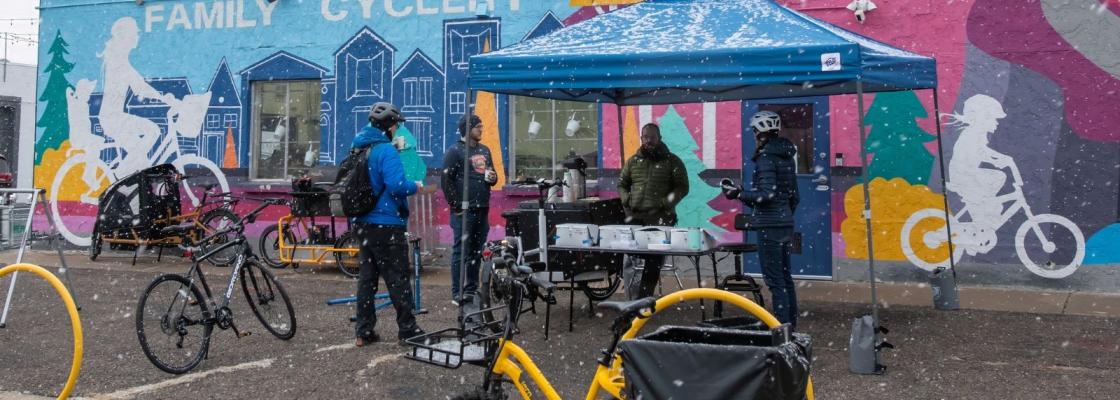 A group of people gathered together under a text with their bicycles surrounding them. They stand in front of a vibrant, colorful mural and there is snow falling from the sky.