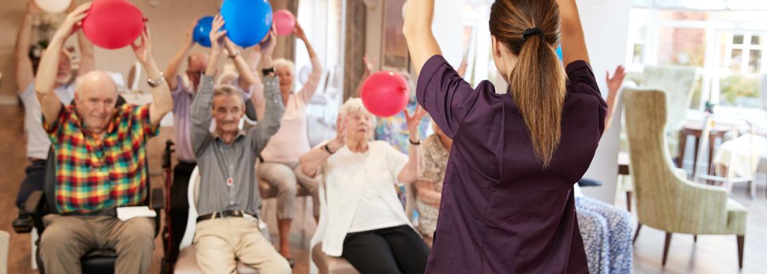 Older adults sitting in a recreational room raising large balls over their heads for daily exercise.