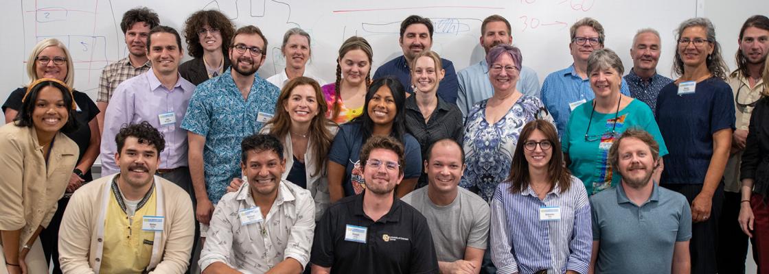 A large group of smiling people posed in three rows in front of a whiteboard.