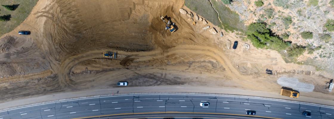 An aerial view of heavy equipment in a large patch of dirt next to cars and trucks driving on a highway.
