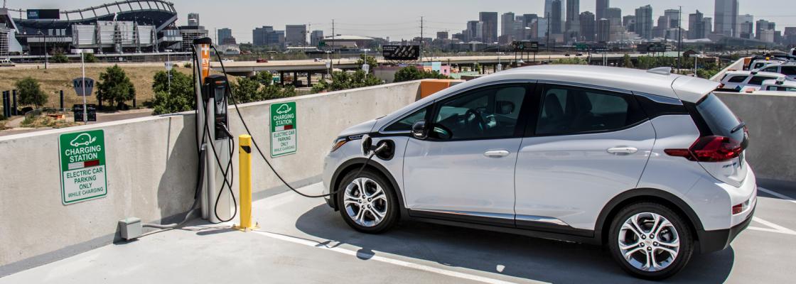 An electric car plugged into a charging station at the top of a parking garage with the Denver city skyline in the background.
