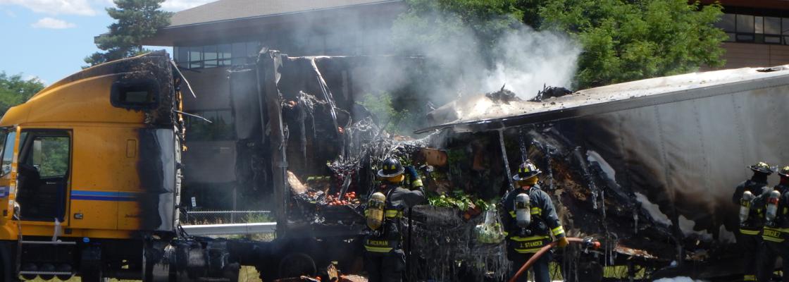 wo firefighters standing on a road in front of the smoking wreckage of a semitruck.