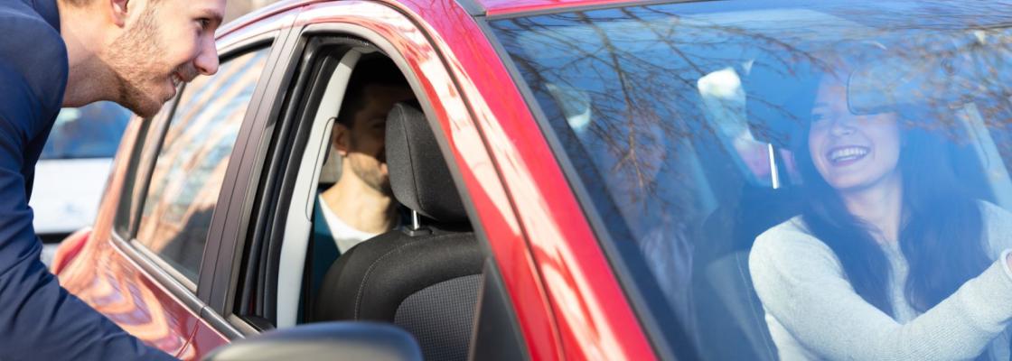 A man reaching to open a car door. There is a person in the driver's seat and two people in the second row of seats.