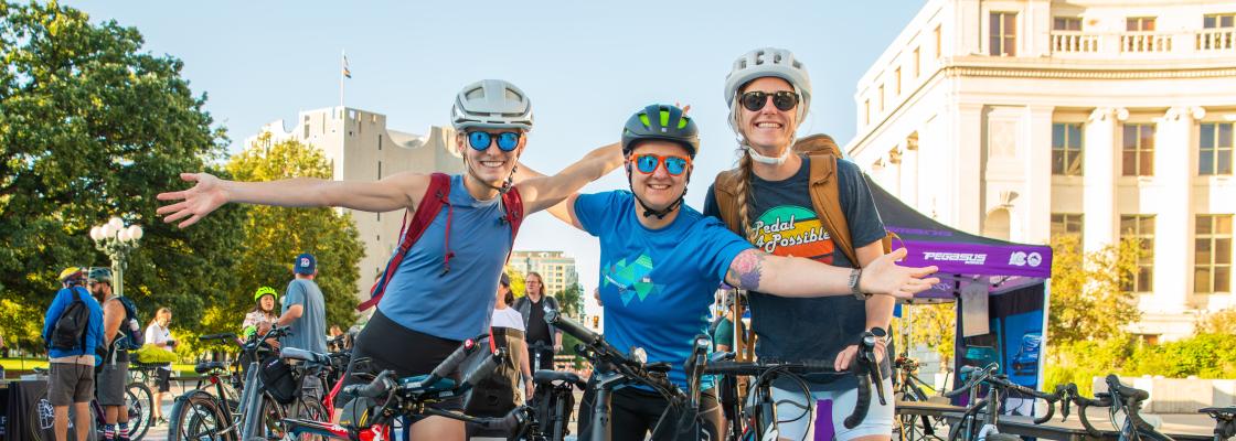Three cyclists wearing helmets smile while posing with their bikes at the 14th and Bannock Bike to Work Day station in downtown Denver.