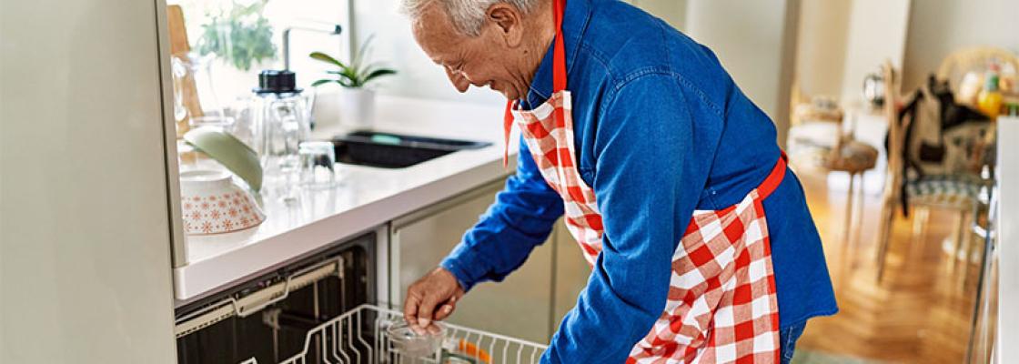 Older adult man smiling while using the dishwasher in the kitchen. 