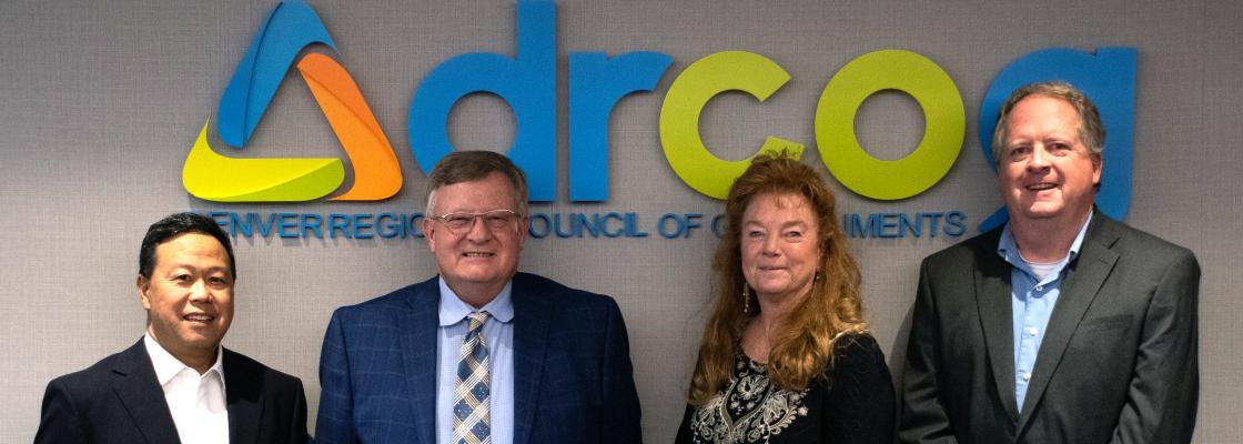 Members of DRCOG's executive committee, all smiling and dressed in professional attire in front of DRCOG's logo, and including a Japanese man with short black hair, a white man with short gray hair and glasses, a white woman with long,red hair and dangly earrings and a white man with short, wavy hair.