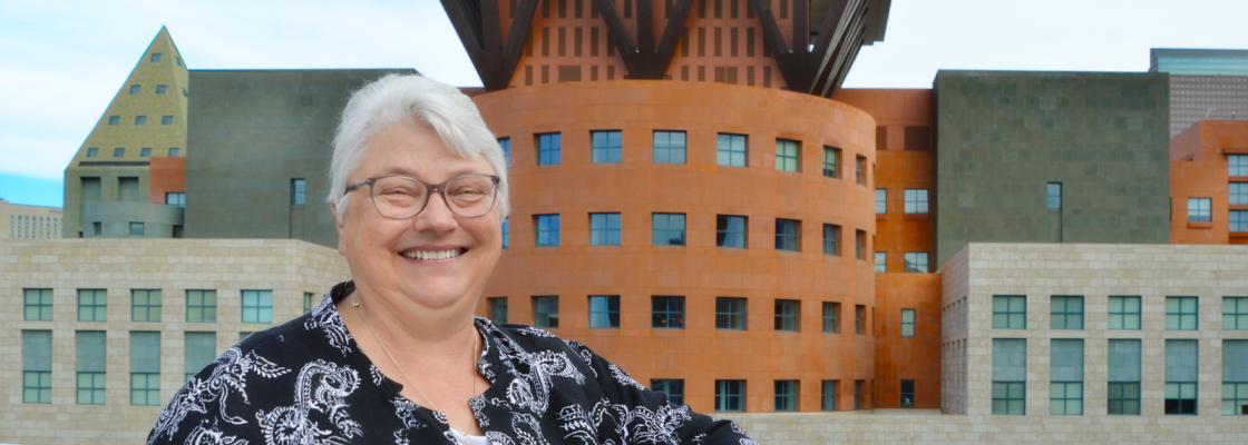 An older white woman wearing glasses smiles. She's standing in front of the Denver skyline.