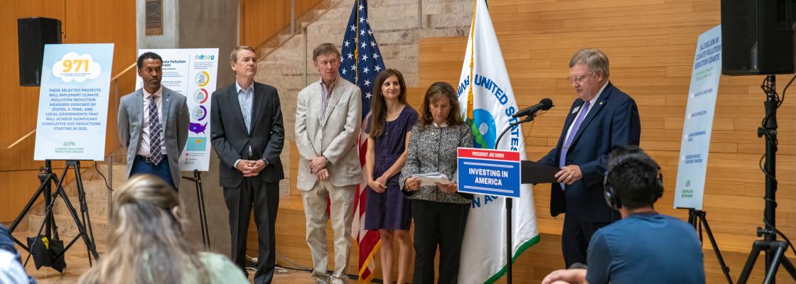 Five people stand in front of flags stand to the left of a man speaking at a lectern.