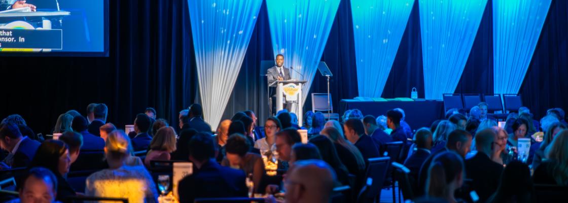 A Black man in a suit addresses a full and festive banquet hall.