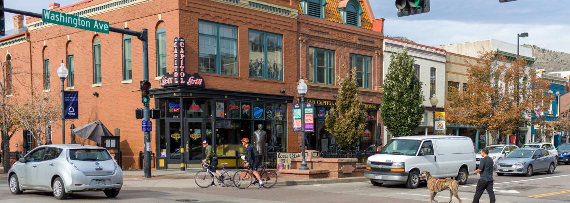 Downtown Golden, Colorado, two people are riding bicycles as a silver car passes them and a man walks a dog through an intersection as cars are stopped.