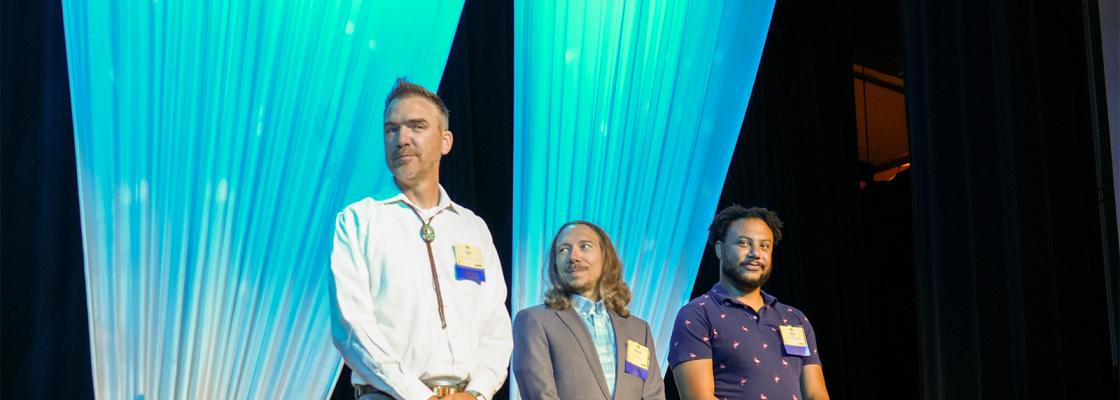 Three men stand on a stage with a blue curtain in the background.