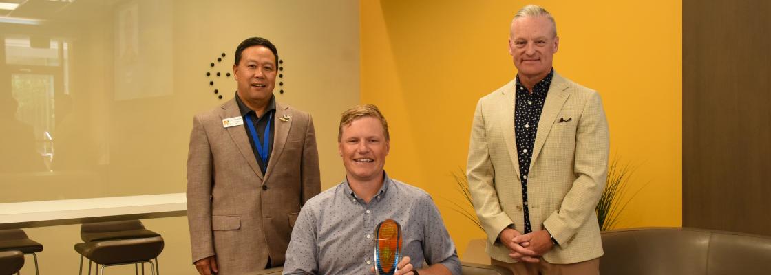 A middled-aged white man sitting holding an award with middle-age Asian appearing man standing to the left and a middle-age white man standing to the right.