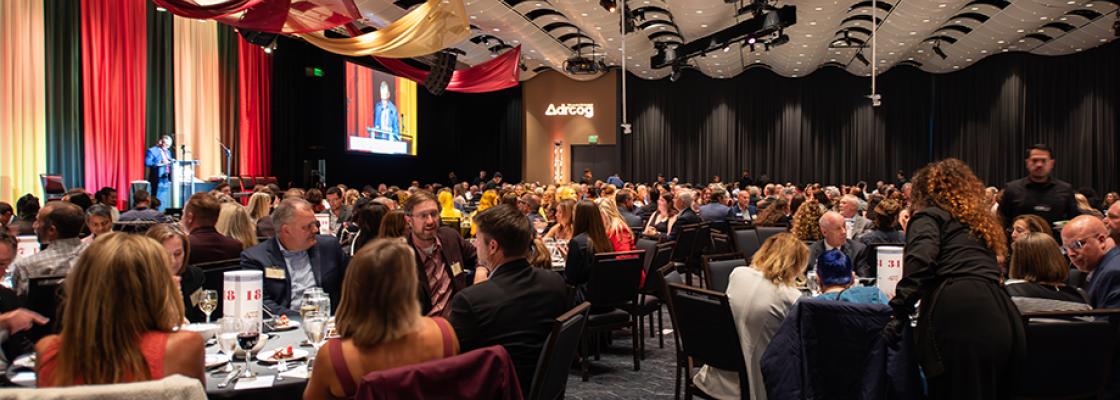 Hundreds of people sit at round tables in a ballroom, listening to a man who is speaking on-stage at the front of the room.