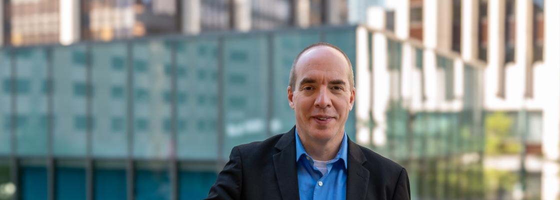 A middle-aged white man wearing a blue ope-collard shirt with a black suit coat standing on a roof-top in downtown Denver.