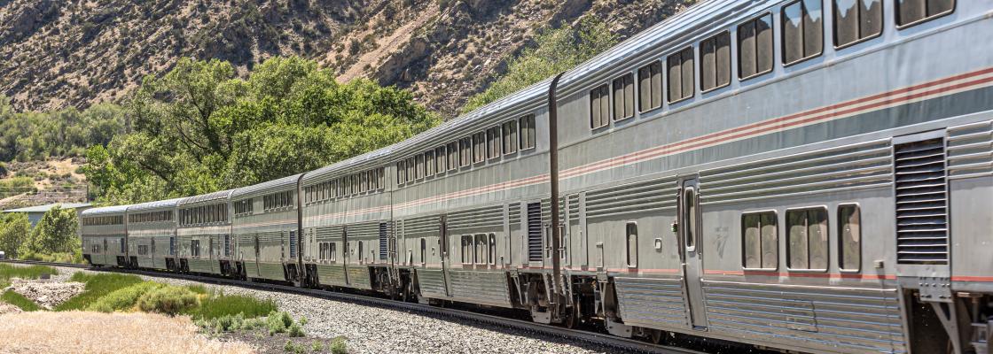 A passenger train travels through a mountain pass with trees in the background.