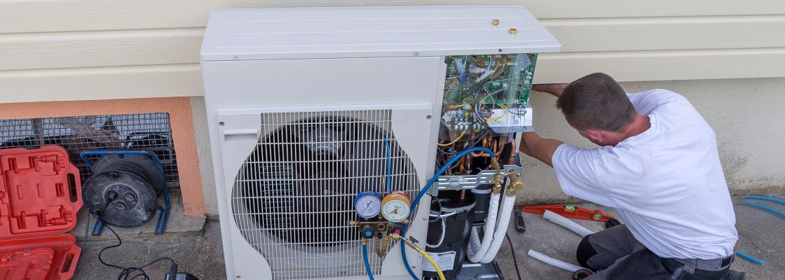 Technician kneeling beside an outdoor HVAC unit, connecting gauges and tools while performing maintenance on exposed system components.