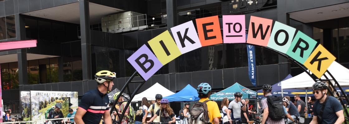 An arch with colored papers spelling out Bike to Work Day in front of an office building in downtown Denver.  A crowd of people wearing bike helmets and riding bikes congregate in front of outdoor tables.