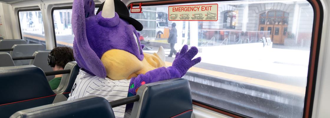 A person in a purple dinosaur costume with horns, resembling the Colorado Rockies' mascot, is seated inside a train car looking out the window at a station platform.