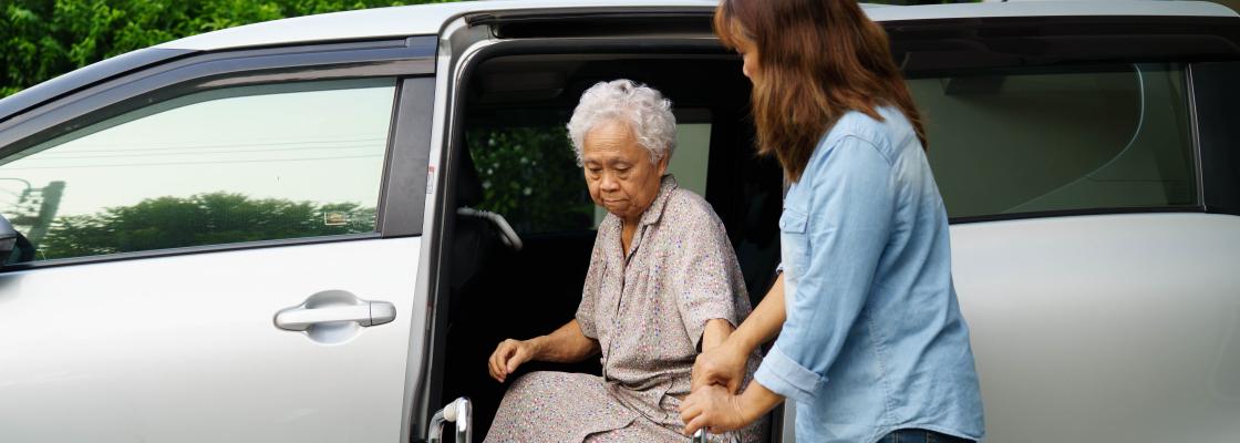 A female caregiver assists and older adult Asian woman who uses a walker with exiting a motor vehicle.