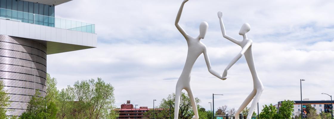 A photo of a park with two giant white sculptures dancing in the middle and buildings in the background.