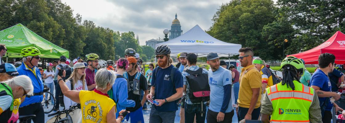 Cyclists gather at a Bike to Work Day station in Civic Center Park, standing around tables with snacks and information booths. People wear helmets and casual or cycling attire, with branded tents (including “Way to Go” and Lime) and the Colorado State Capitol dome visible in the background under a cloudy sky.