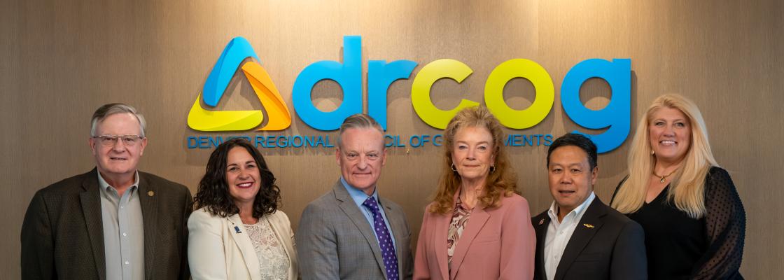 Six middle-aged people pose for a group photo in front of a Denver Regional Council of Governments wall logo inside an office.