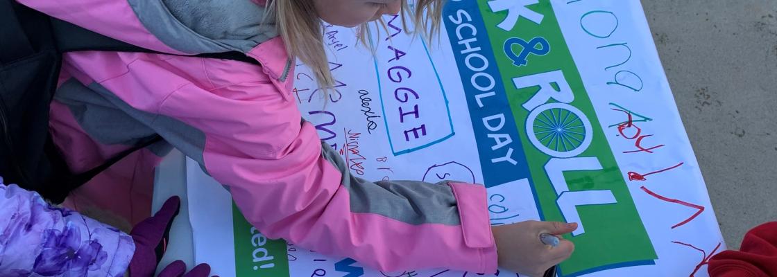 Children gather around a table signing a Walk & Roll to School Day banner with colorful markers.