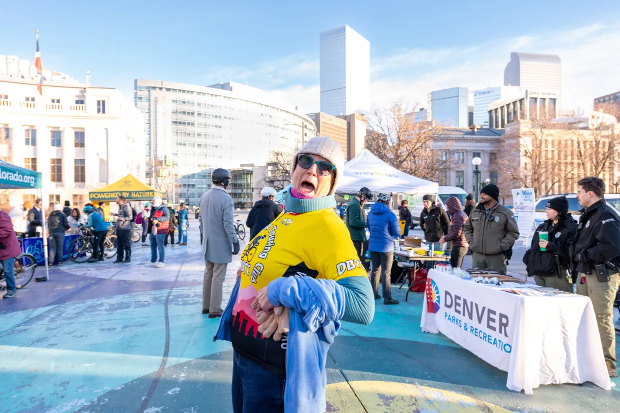 An early morning view of the downtown Denver skyline from the bike lane in front of the Denver City Council building. A smiling cyclist stands at the foreground and other smiling people hold and ride their bikes while socializing at tables hosted by various vendors.