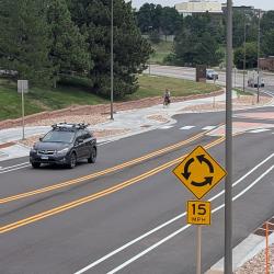 A black car drives down a mostly empty road with bike lanes and new sidewalks on each side. A small roundabout can be seen in the background.