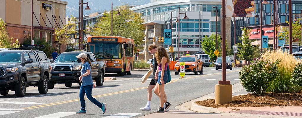 Three people walk in the crosswalk of a busy intersection in Boulder, CO.